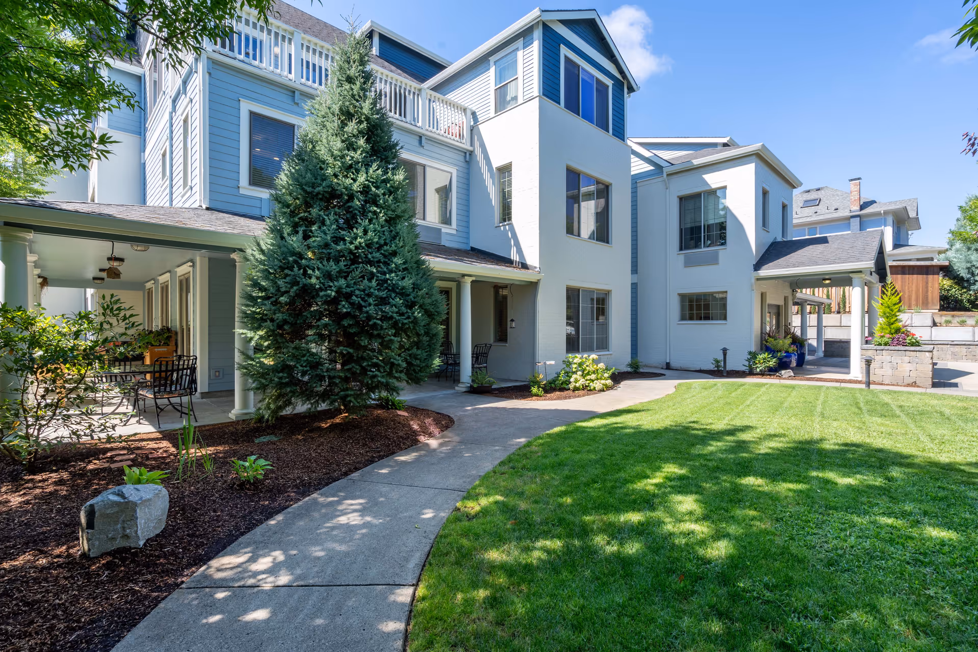 Exterior view of a senior living community building with a well-maintained lawn, a curved concrete walkway, and a large evergreen tree near the entrance. The building has multiple windows and a covered porch area with outdoor seating.