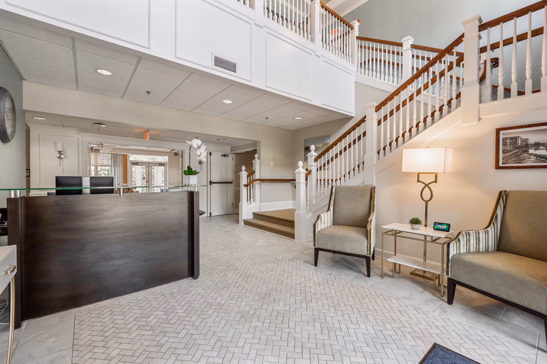 A bright and spacious reception area with a dark wooden front desk, two upholstered chairs with striped armrests, a small side table with a lamp and a digital clock, and a staircase with white railings and wooden handrails leading to an upper floor.