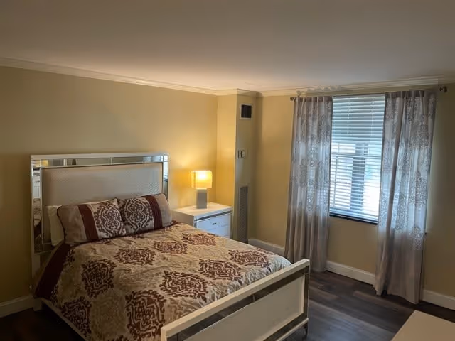 A bedroom with a bed featuring a patterned bedspread and pillows, a nightstand with a lamp, beige walls, a window with blinds and sheer curtains, and dark wood flooring.