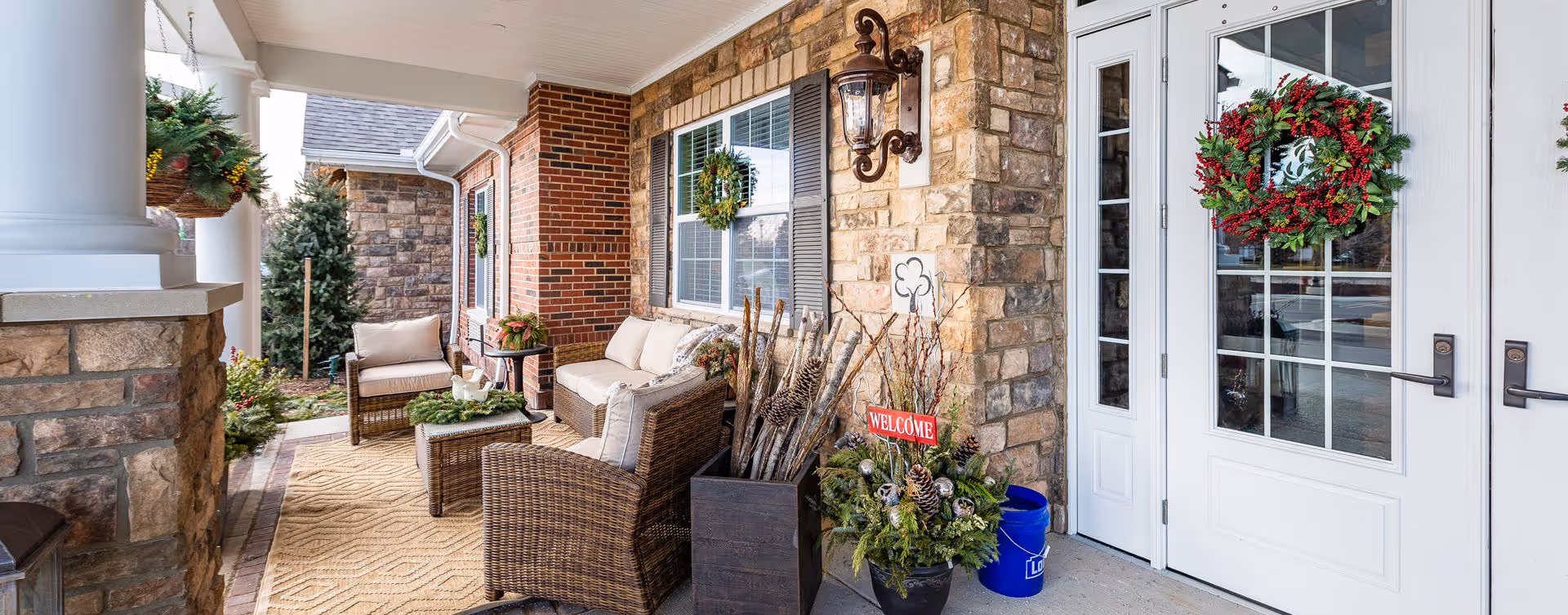 Covered outdoor porch area with wicker seating including chairs and a sofa with cushions, a small table, and decorative plants. The porch has stone and brick walls, a hanging lantern light fixture, and two white doors with glass panes adorned with festive wreaths. There is a welcome sign among the plants near the entrance.