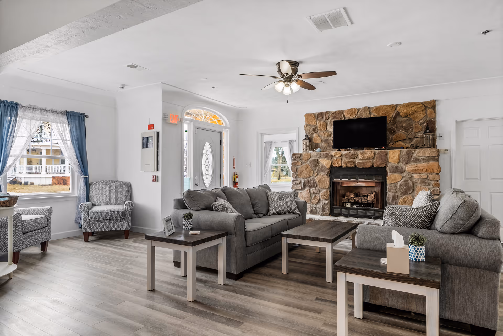 A bright and spacious living room in an assisted living facility featuring two gray sofas with patterned cushions, two gray armchairs near a window with blue curtains, a stone fireplace with a mounted flat-screen TV above it, and several wooden coffee and side tables. The room has light wood flooring, white walls, a ceiling fan with lights, and a glass front door with sidelights.