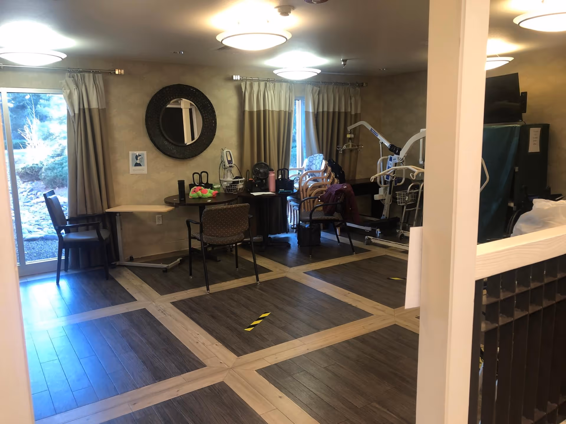 A communal activity room with chairs and tables, medical lift equipment, a round mirror on the wall, and large windows.