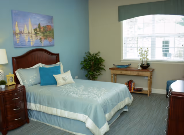 A neatly made bed with light blue bedding and decorative pillows in a senior living bedroom. The room features a wooden nightstand with a lamp, clock, and small plant. There is a painting of sailboats on the wall above the bed, a green potted plant in the corner, a wooden table with books and decorative items under a large window with blinds, and a dresser on the right side.