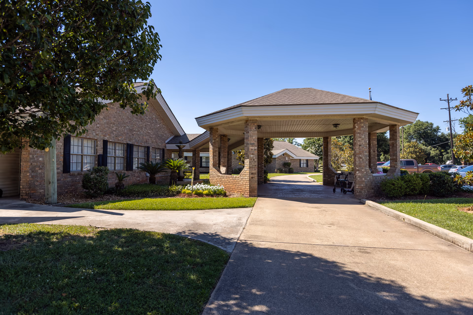 Exterior view of a senior living facility with a covered driveway supported by brick columns. The building is made of brick with black window shutters and surrounded by green grass and trees under a clear blue sky.