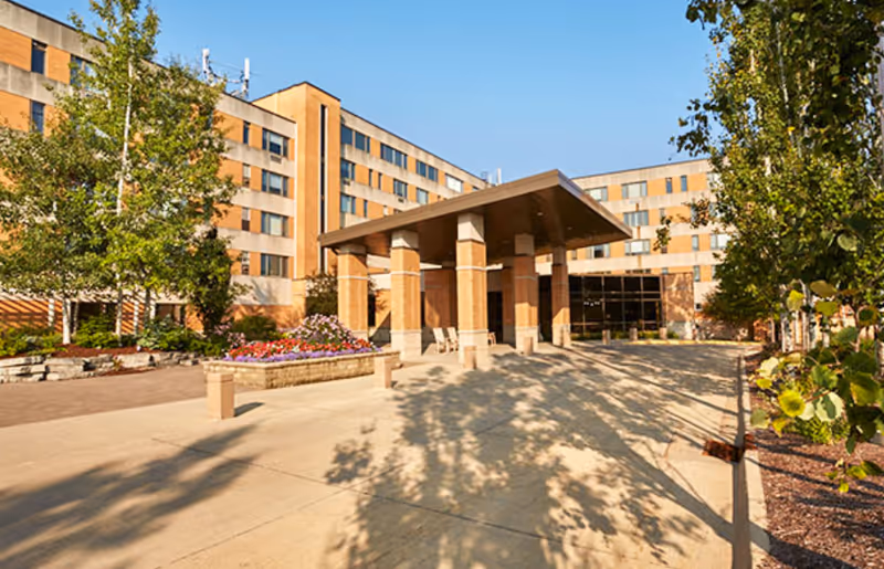 Exterior view of a multi-story senior living facility with a covered entrance supported by brick columns, surrounded by trees and flower beds under a clear blue sky.