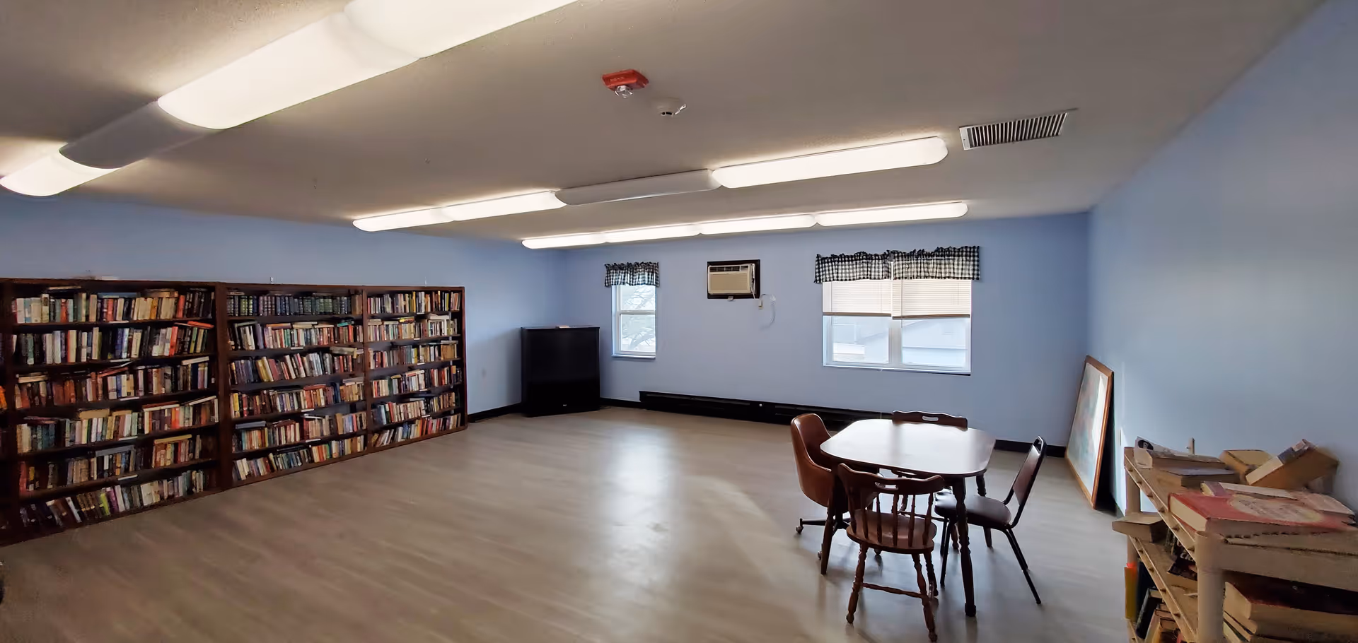 Spacious senior community activity room with bookshelves along one wall, a small table and chairs, windows, and overhead fluorescent lights.