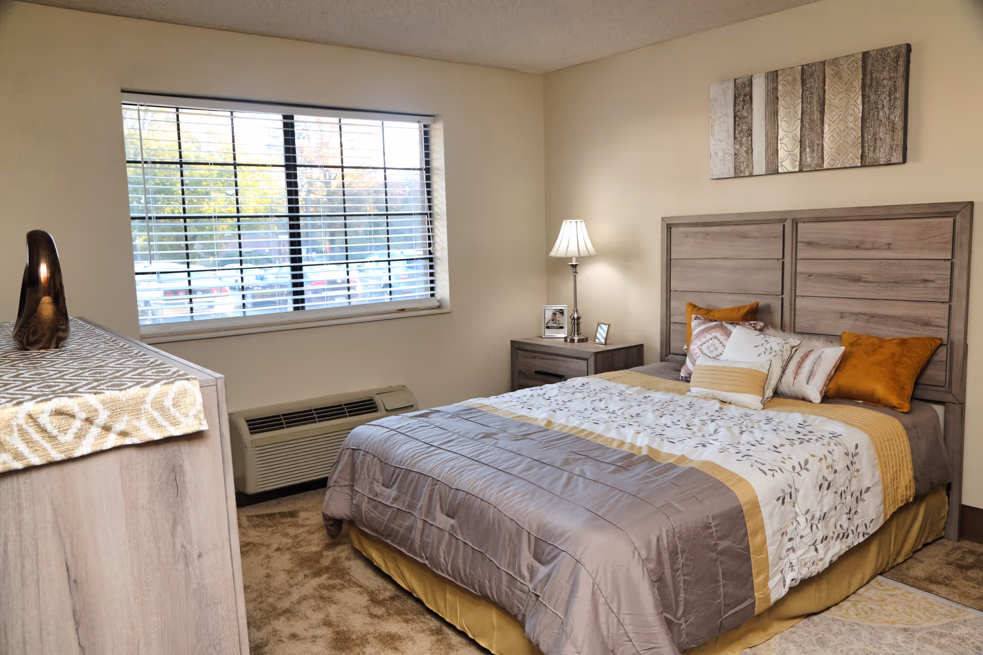 Bright furnished bedroom with a made bed, wooden headboard, nightstand with lamp, dresser, and a window with blinds.