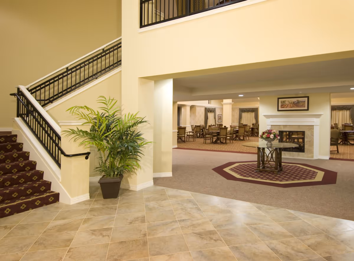 Bright interior lobby of an assisted living facility with a staircase, potted plant, tiled floor, and a seating/dining area with a fireplace and a table with flowers.
