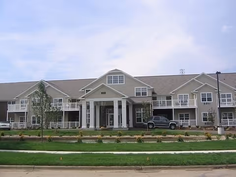 Two-story beige senior living building with a covered central entrance, balconies, and landscaped lawn in front.
