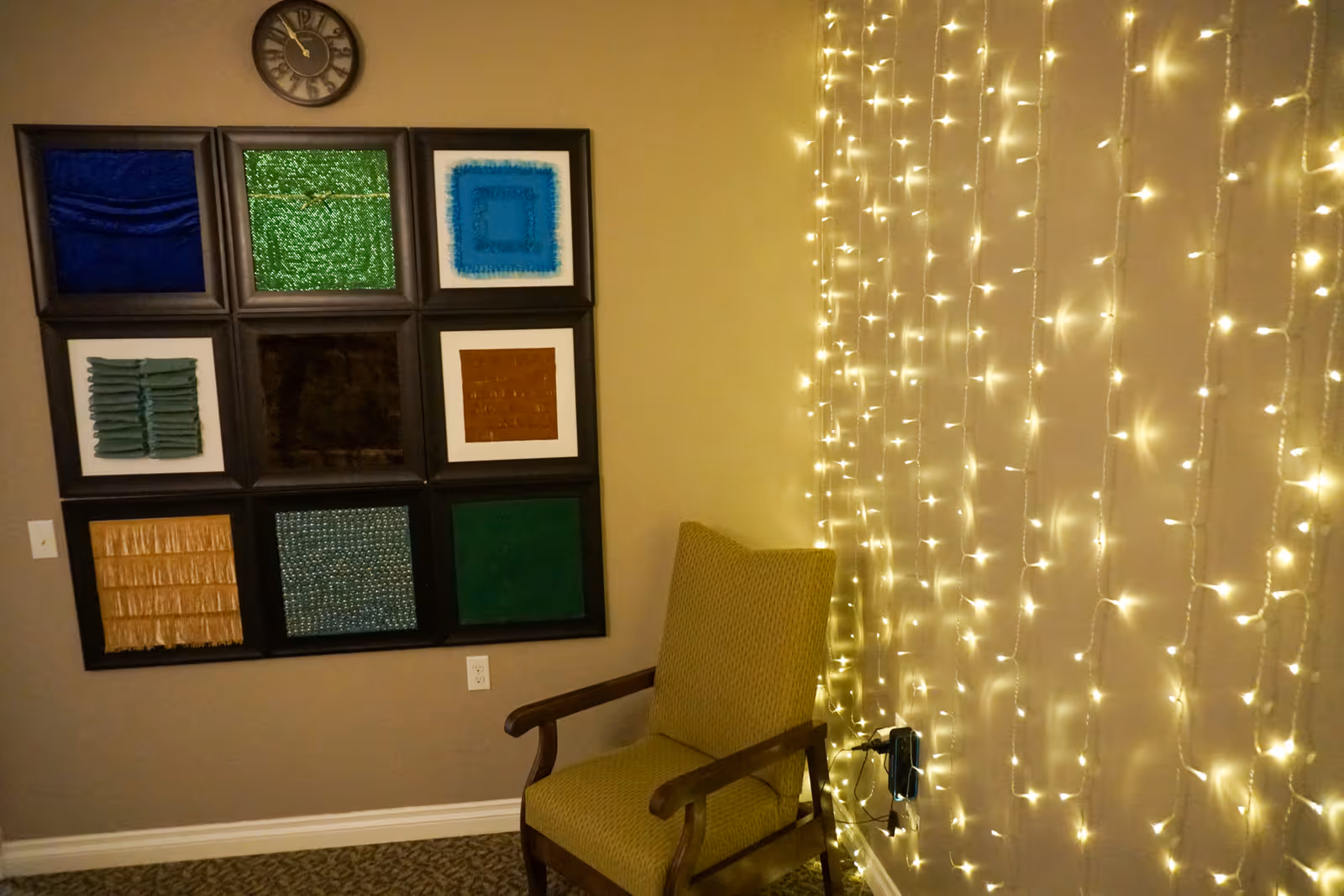 A cozy corner of a room with a green upholstered armchair next to a wall decorated with hanging string lights. On the adjacent wall, there is a large framed art piece composed of nine square sections with different textured and colored materials. A round clock is mounted above the artwork.