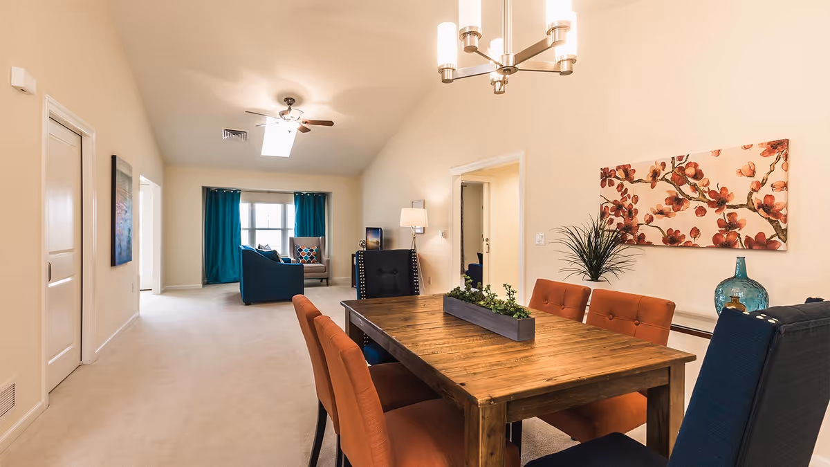 Open-plan living and dining area with a wooden dining table surrounded by colorful upholstered chairs and a sitting area with teal curtains in the background.