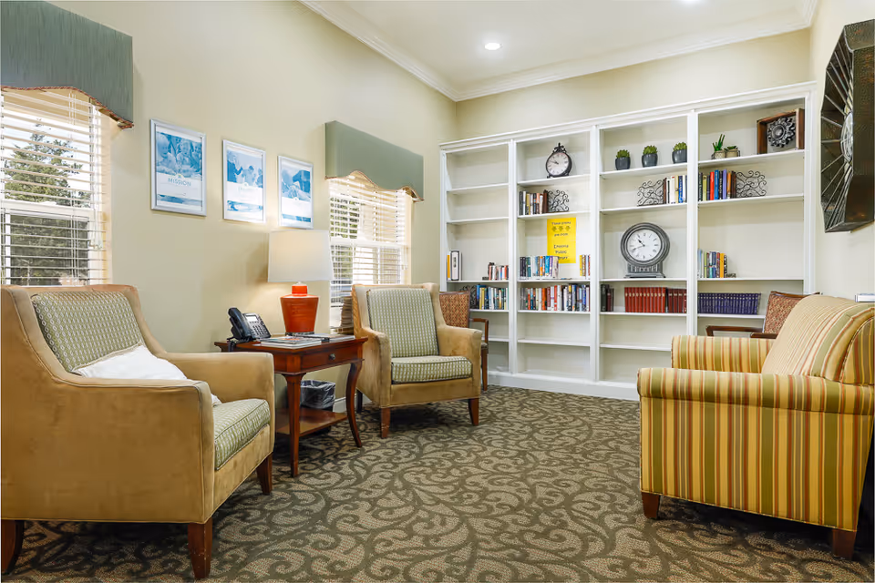 A cozy sitting area in an assisted living facility with three upholstered chairs arranged around a wooden side table with a lamp and a phone. Behind the chairs is a large white bookshelf filled with books, clocks, and small potted plants. The room has two windows with blinds and green valances, light-colored walls, and patterned carpet flooring.