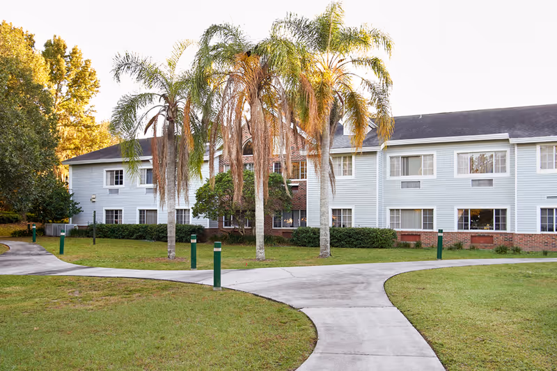 Front exterior of a two-story senior living building with palm trees and curved concrete walkways across a grassy lawn.