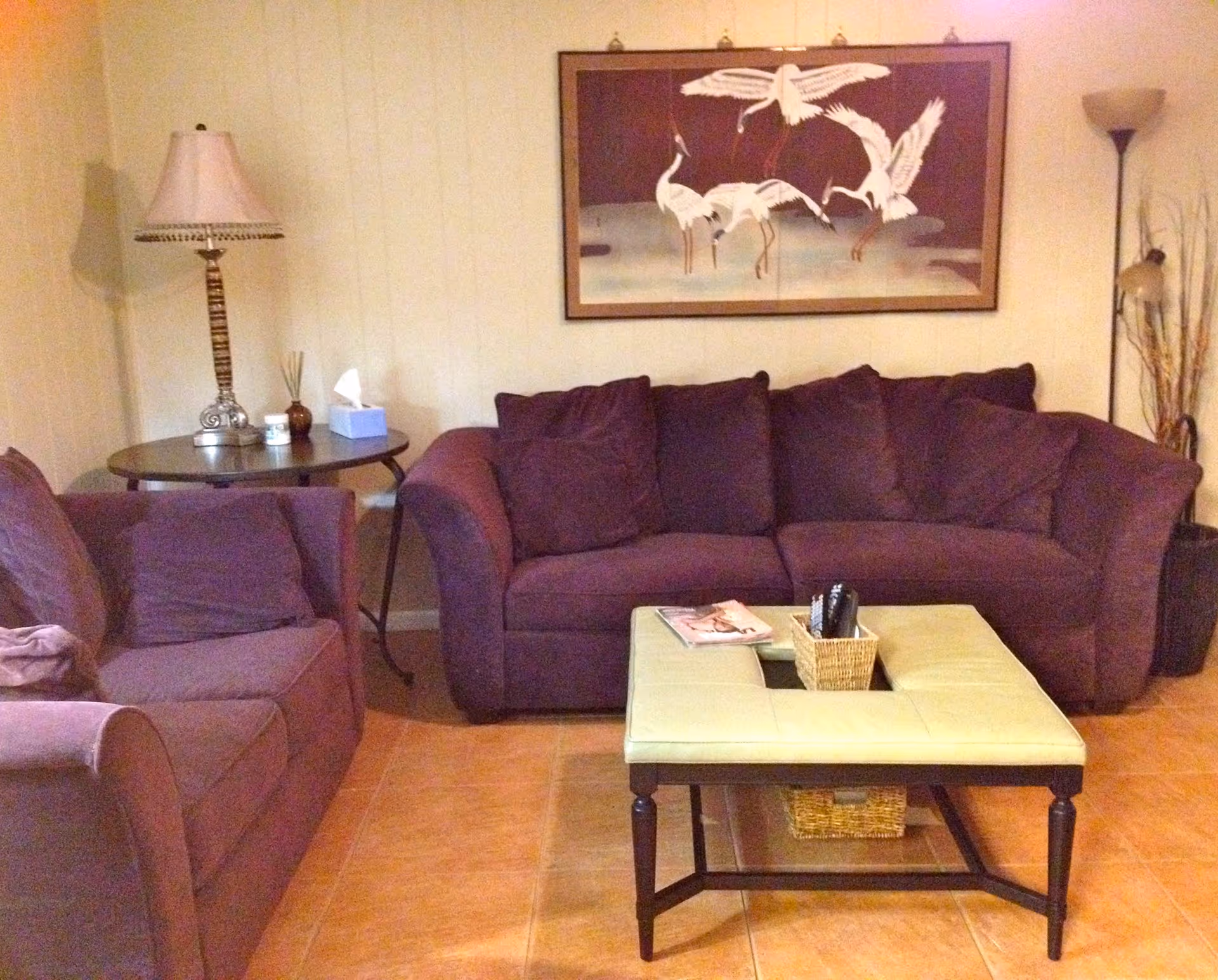 A cozy living room with two purple upholstered sofas, a square green ottoman coffee table with a wicker basket holding remote controls and a magazine on top. There is a round side table with a decorative lamp, tissue box, and small items. A tall floor lamp and decorative dried plants are in the corner. A framed artwork of white cranes is hanging on the wall behind the sofa.