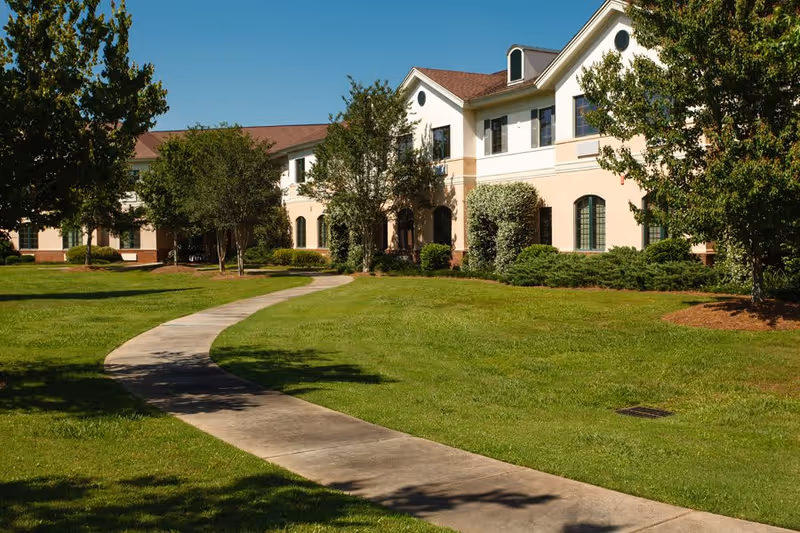 A sunny outdoor view of a senior living facility with a curved concrete walkway leading through a well-maintained grassy lawn. The building has two stories with beige and white walls, multiple windows, and a brown roof. Trees and shrubs surround the building and walkway under a clear blue sky.