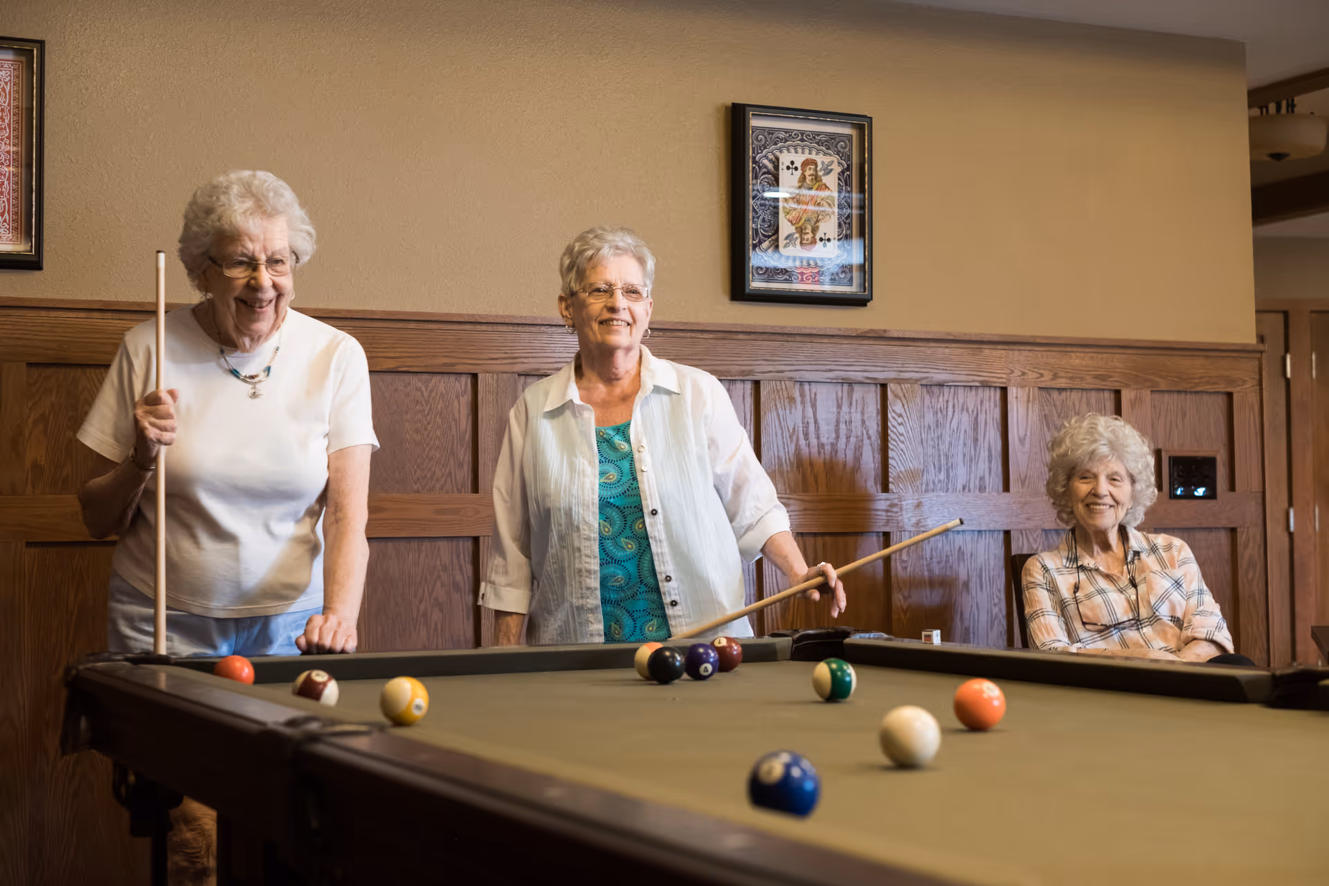 Three elderly women enjoying a game of pool in a room with wooden paneling and framed artwork on the wall. Two women are standing and holding pool cues, while the third woman is sitting and smiling.