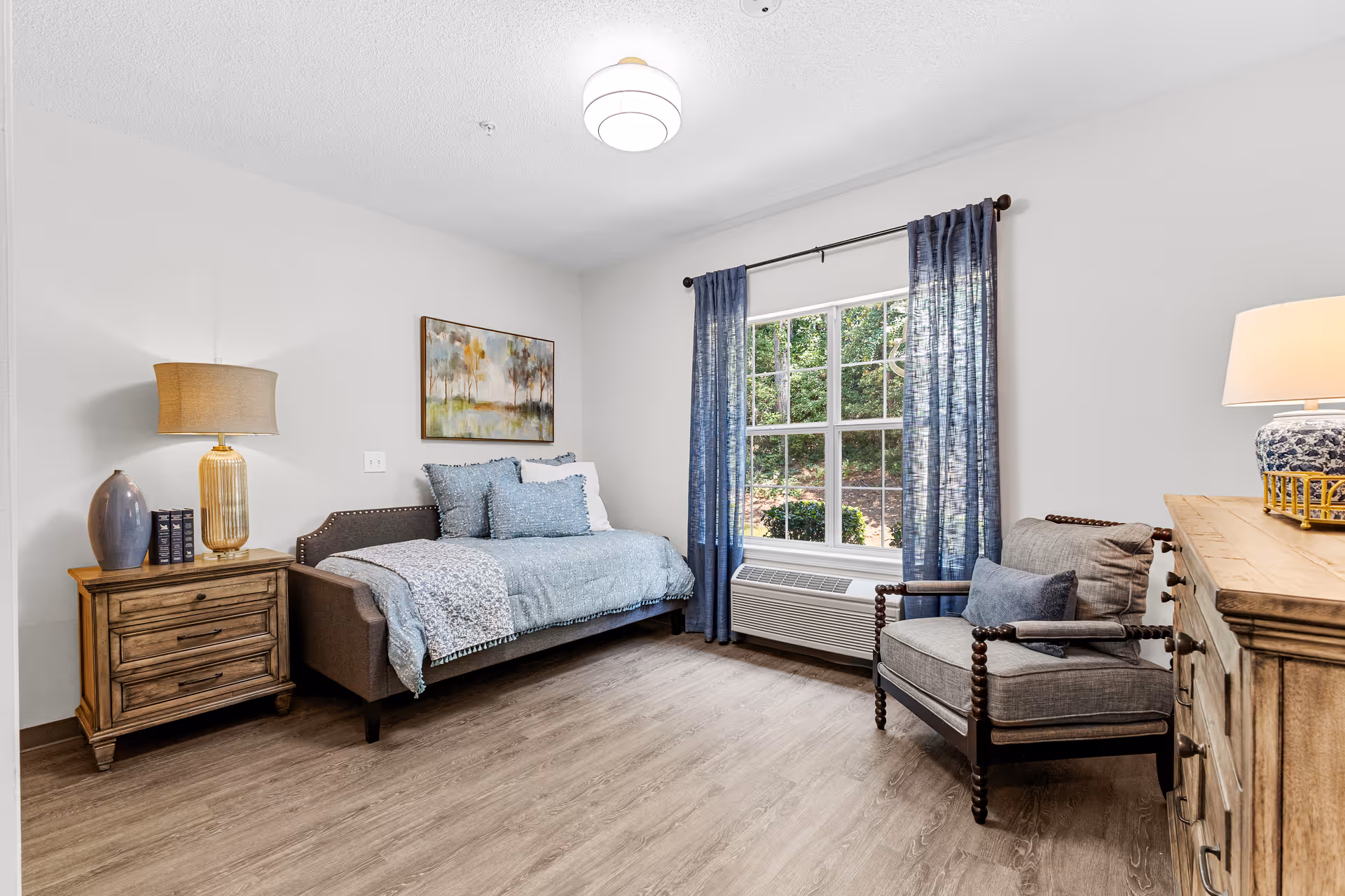 A cozy bedroom with a daybed covered in blue bedding and pillows, a wooden nightstand with a lamp and decorative items, a window with blue curtains showing greenery outside, a cushioned armchair with a pillow, and a wooden dresser with a lamp on top.