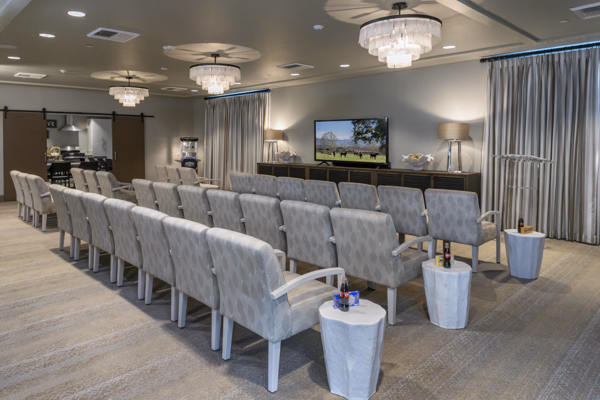 Rows of upholstered chairs facing a wall-mounted TV in a carpeted community screening room with chandeliers and side tables.