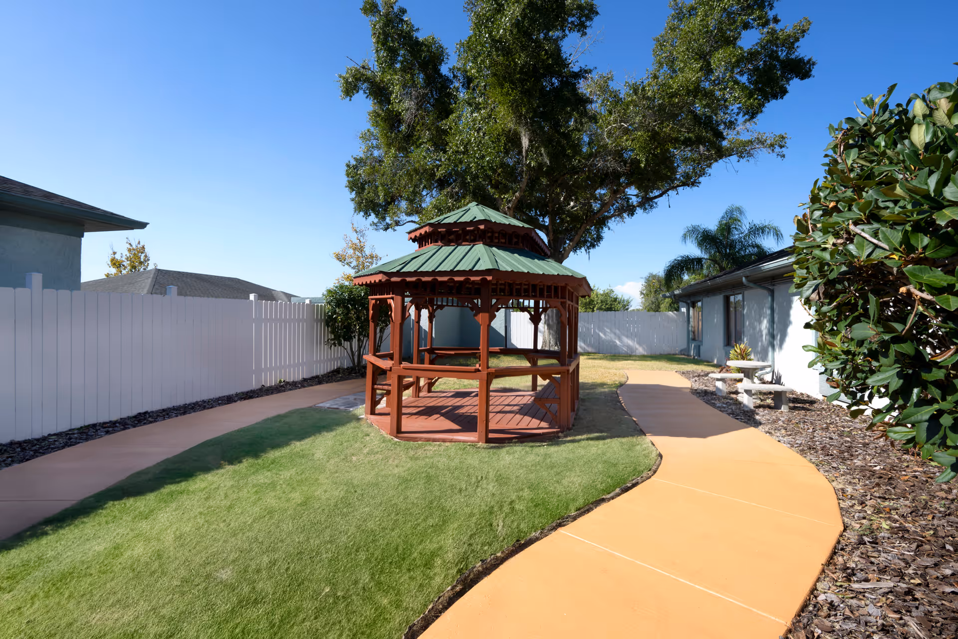 Outdoor garden area with a wooden gazebo featuring a green roof, surrounded by a curved paved walkway, green grass, bushes, and a white fence under a clear blue sky.