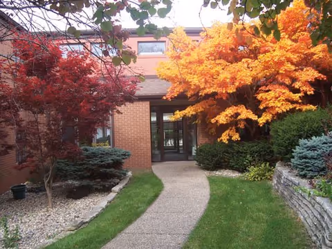 Pathway leading to the entrance of a brick building surrounded by colorful autumn trees and well-maintained landscaping with green grass and shrubs.