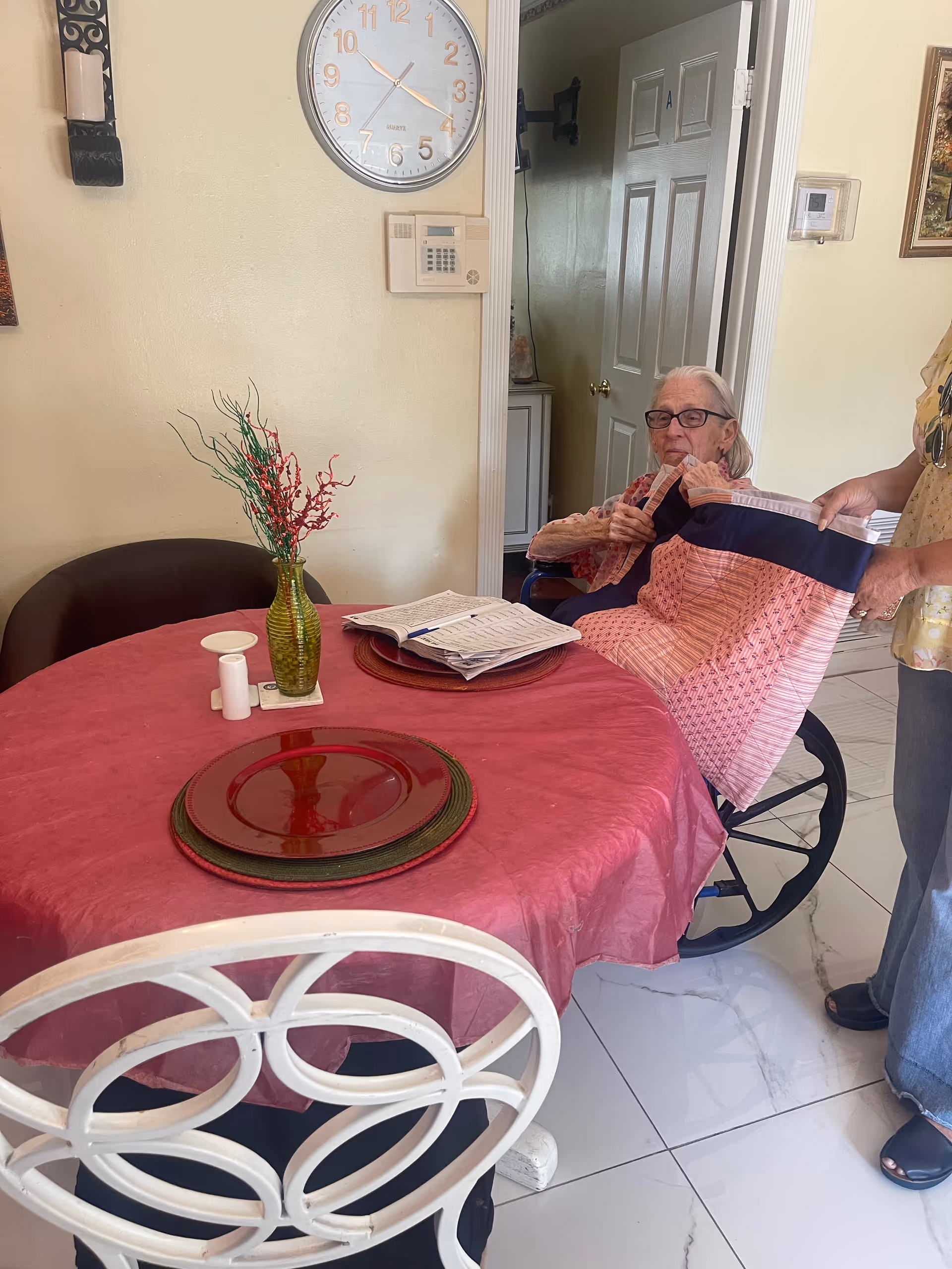 An elderly woman in a wheelchair is seated at a round dining table covered with a red tablecloth. A person standing next to her is helping adjust a pink and navy blue garment on her. The table has a green vase with red and green decorative branches, a white candle holder, and a stack of papers or a book. A large wall clock and a security keypad are visible on the wall behind them.