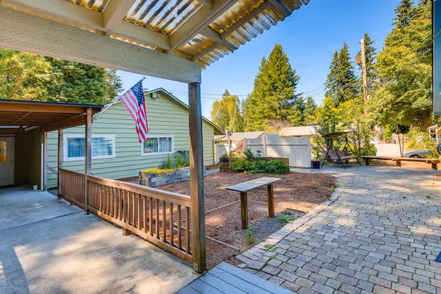 Outdoor view of a senior living facility showing a covered porch with wooden railing, an American flag hanging from the porch, a paved walkway, a garden area with mulch and plants, a swing bench, and several trees and shrubs under a clear blue sky.