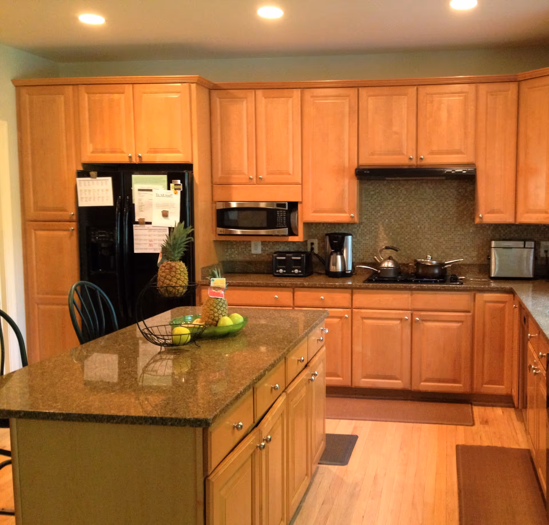 A kitchen with wooden cabinets and a granite countertop island. The kitchen features a black refrigerator with papers attached, a microwave, a toaster, a coffee maker, a stove with pots, and a fruit basket with pineapples and apples on the island. The floor is wooden and there are two chairs partially visible.
