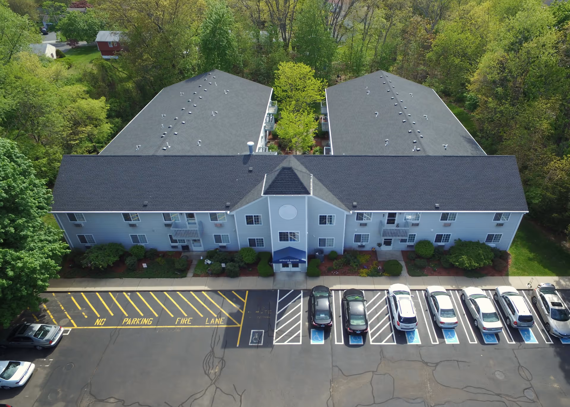 Aerial view of Spruce Manor, showing a two-story residential building with a dark roof, surrounded by trees and greenery. The front of the building has a blue awning over the entrance with the name Spruce Manor. In front of the building is a parking lot with several cars parked, including designated handicapped parking spaces and a marked fire lane.