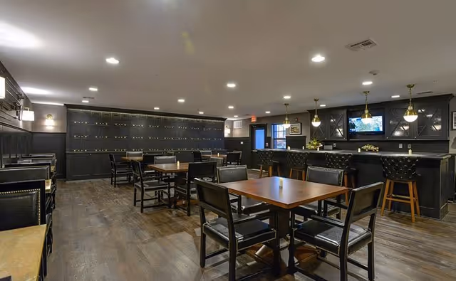 Interior view of a senior living facility dining area with multiple wooden tables and black cushioned chairs arranged neatly on a dark wood floor. The room features a bar counter with high stools, pendant lighting, and a television mounted on the wall above the bar. The walls and cabinetry are painted dark, creating a cozy and elegant atmosphere.