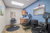 A small salon room with a black salon chair on a black mat, two black salon hair dryers, a black waiting chair, a wooden cabinet with a sink, a wall-mounted mirror, and a skylight providing natural light.