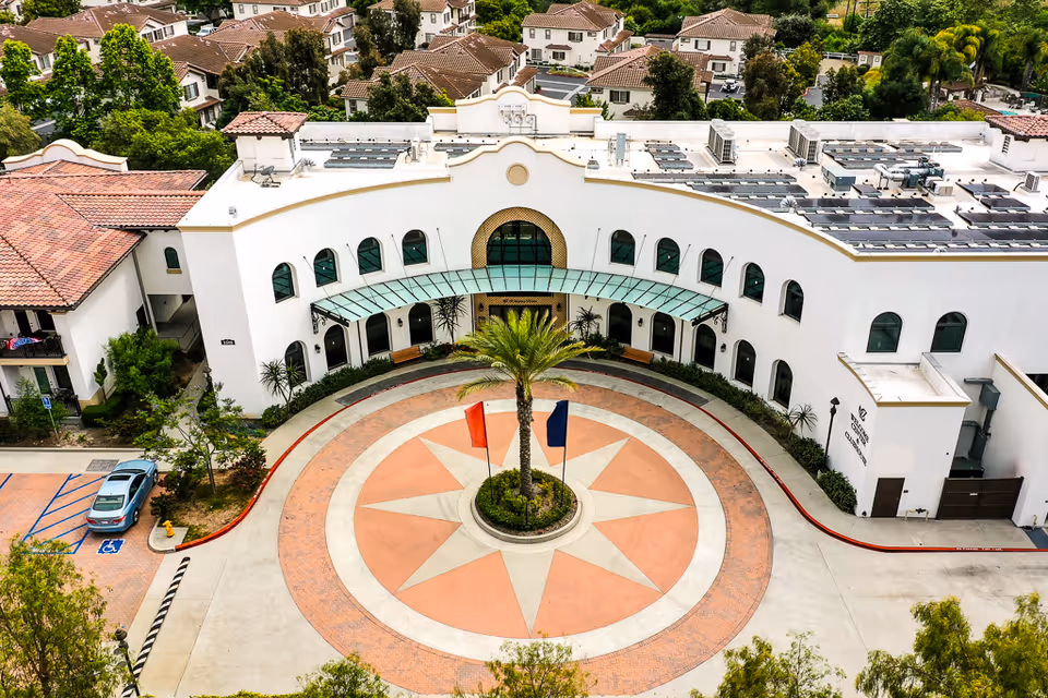 Aerial view of the front exterior of a senior living facility with a circular driveway featuring a star design and two flags in the center. The building has white walls, arched windows, a glass canopy over the entrance, and solar panels on the roof. Surrounding the facility are residential houses and greenery.