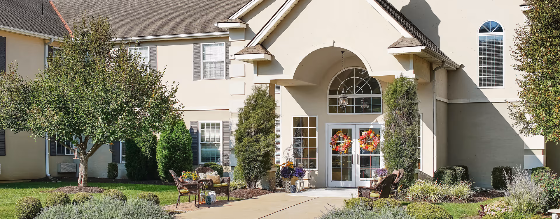 Front entrance of a senior living facility with double glass doors decorated with colorful wreaths. The building has beige walls, large windows with shutters, and a peaked roof. There are two seating areas with wicker chairs and small tables on either side of the entrance, surrounded by well-maintained landscaping including trees, shrubs, and flowering plants.