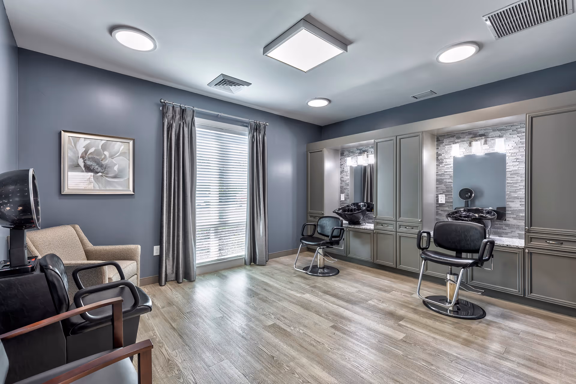 A modern salon area with two black salon chairs in front of mirrors and sinks for hair washing. The room has gray walls, wood flooring, a large window with gray curtains, and additional seating including a beige armchair and a black chair with wooden armrests. The lighting is bright with ceiling fixtures.