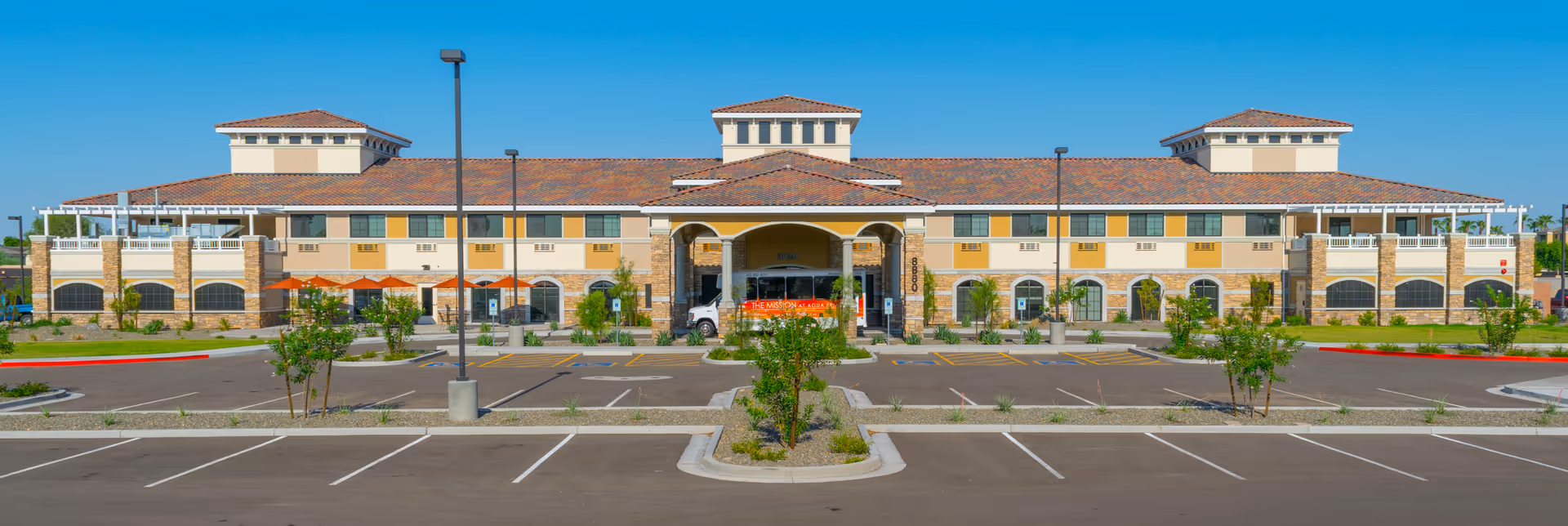 Wide frontal view of a two-story senior living building with a tiled roof, covered main entrance and an empty parking lot in front.
