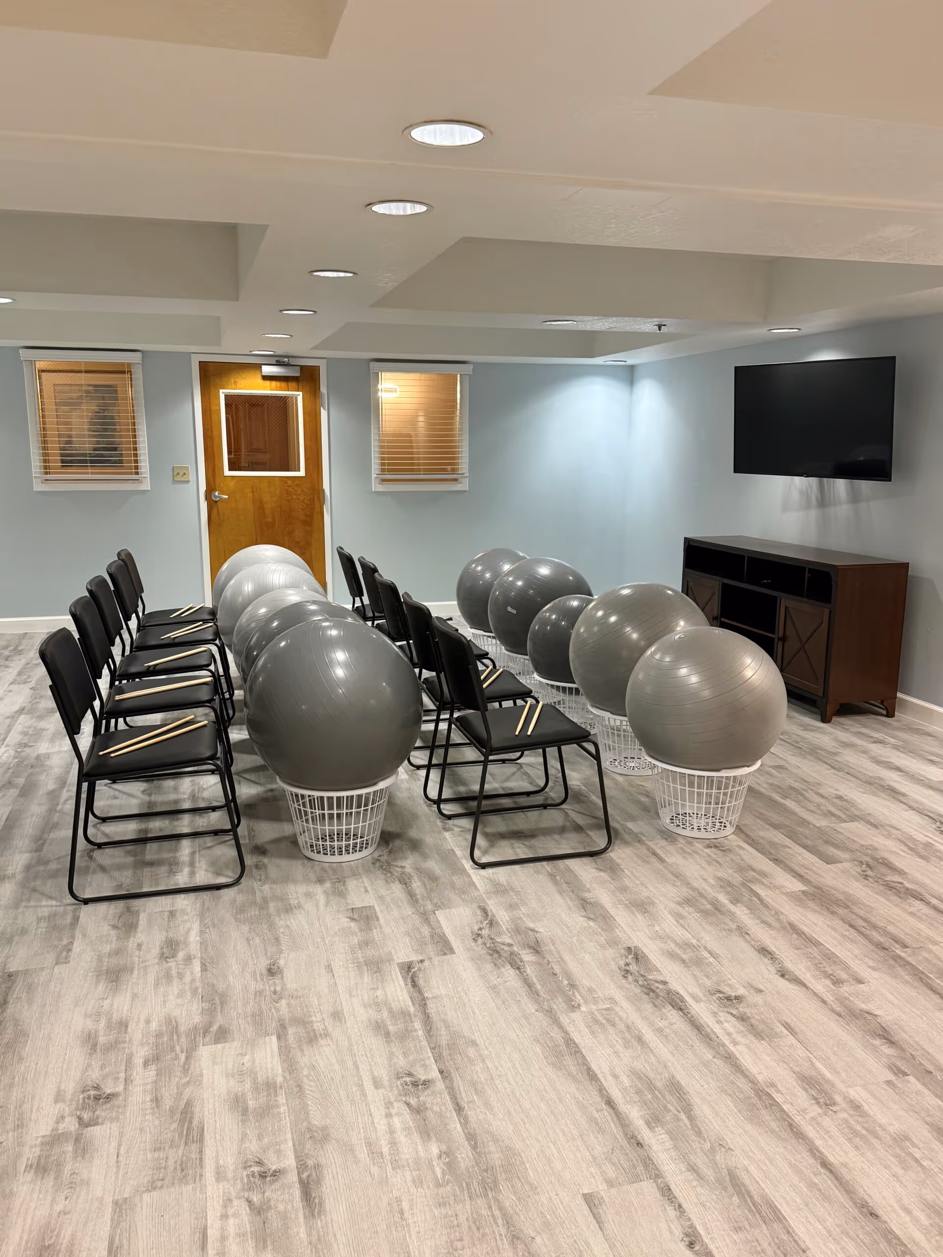 A room with two rows of black chairs facing a wall-mounted flat screen TV. Each chair has a pair of drumsticks on the seat, and in front of each chair is a large gray exercise ball placed on a white basket. The room has light-colored wood flooring, light blue walls, two small windows with blinds, and a wooden door.