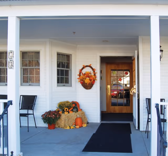 Entrance of a white building with a wooden door featuring glass panels. The porch is decorated with autumn-themed items including a basket with fall leaves, hay bales, pumpkins, and potted flowers. Two black chairs are placed on either side of the porch.