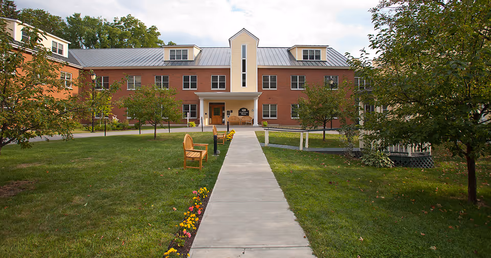 A brick senior living facility building with a metal roof, surrounded by green grass, small trees, and flower beds. A concrete walkway leads to the main entrance, which has a small covered porch with benches on either side. There is a wooden bench along the walkway and a white gazebo visible on the right side.