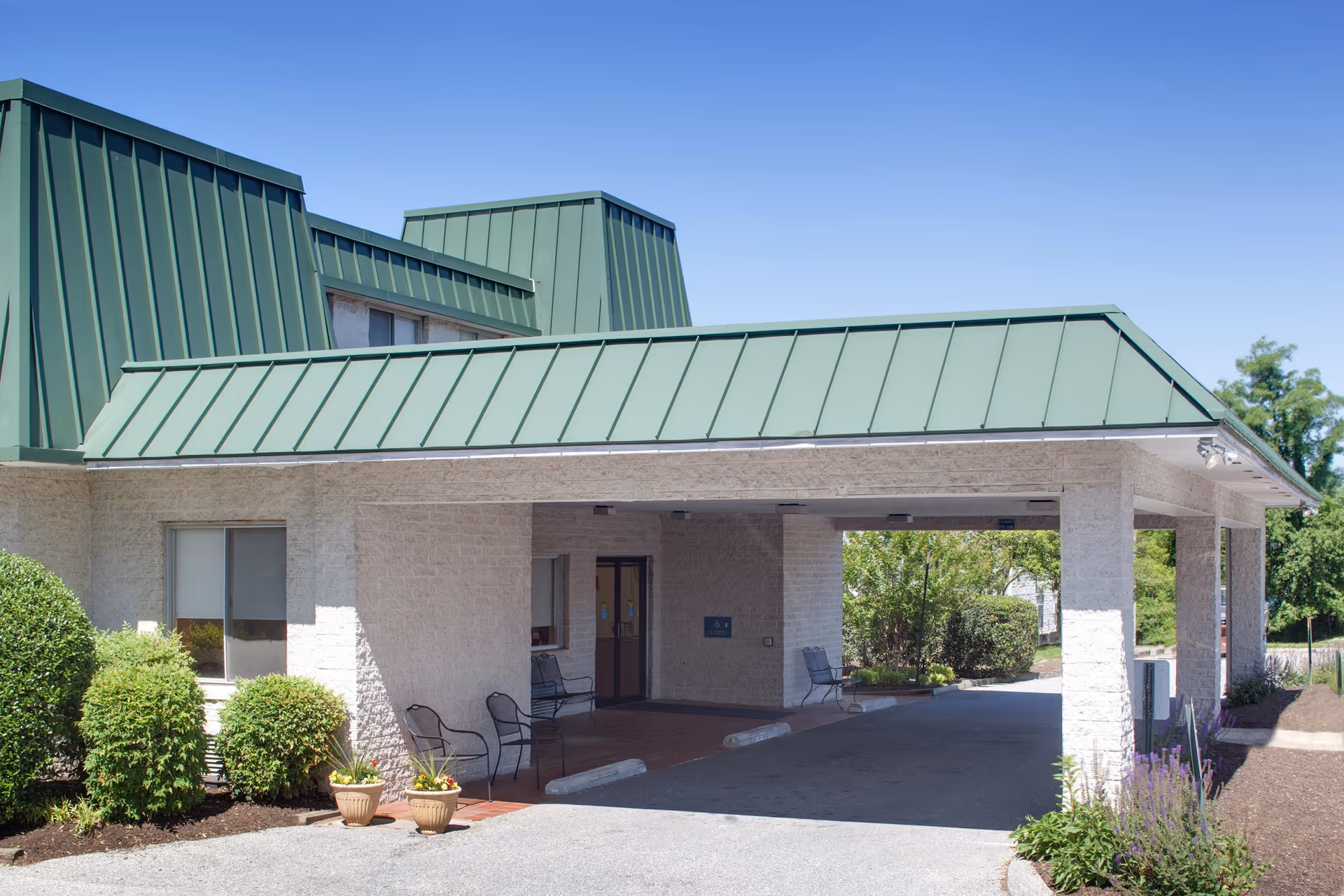 Front entrance of a light-colored brick building with a green metal canopy, outdoor chairs, potted plants, and landscaping.