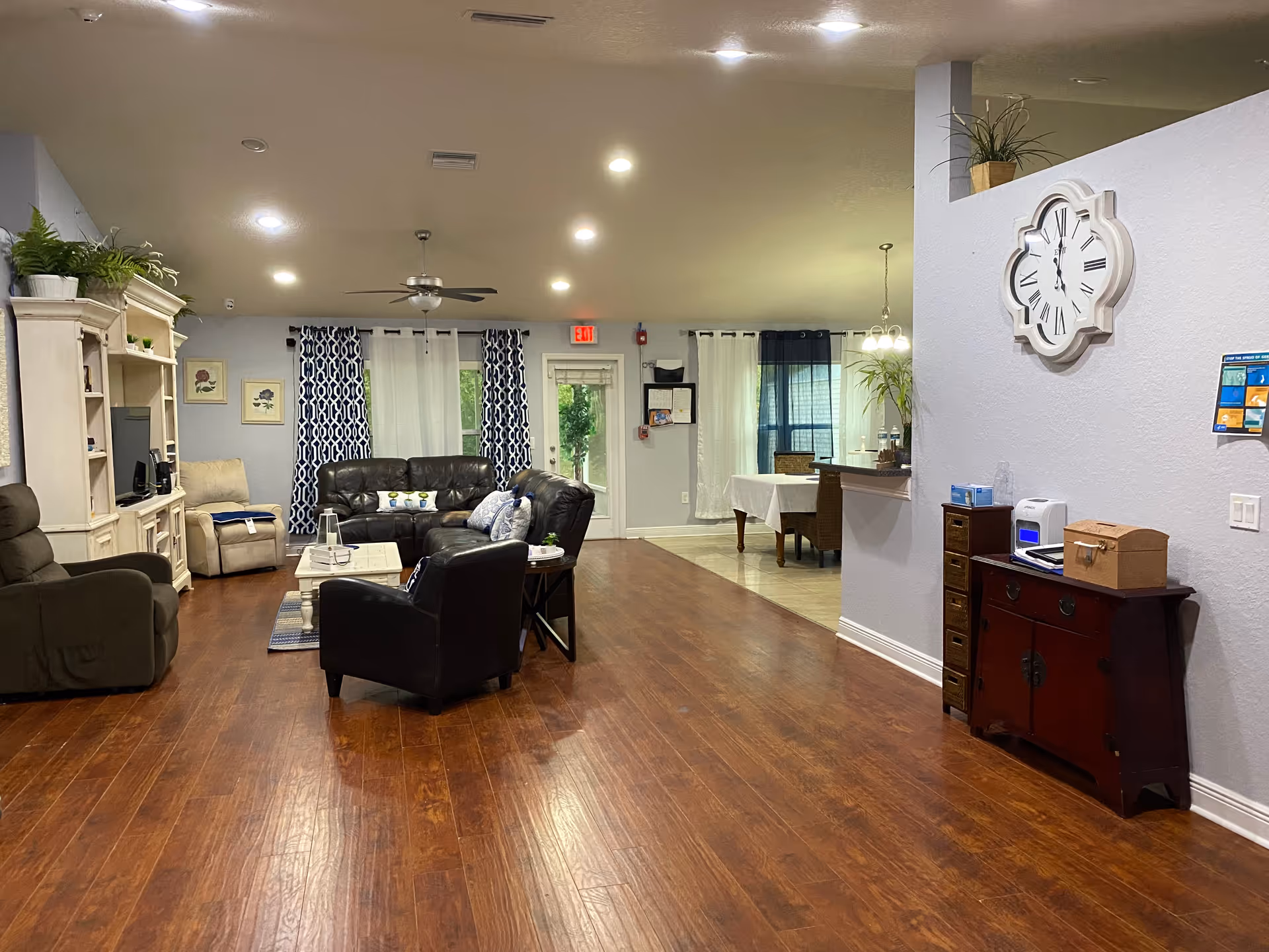 A spacious and well-lit living room area in Mariner Palms Assisted Living Facility featuring wooden flooring, multiple seating options including a black leather sofa, armchairs, and a beige recliner. The room has a white entertainment center with a TV, decorative plants, patterned curtains on the windows, and a ceiling fan. To the right, there is a small wooden cabinet with various items on top and a large decorative wall clock above it. In the background, a dining area with a table and chairs is visible.