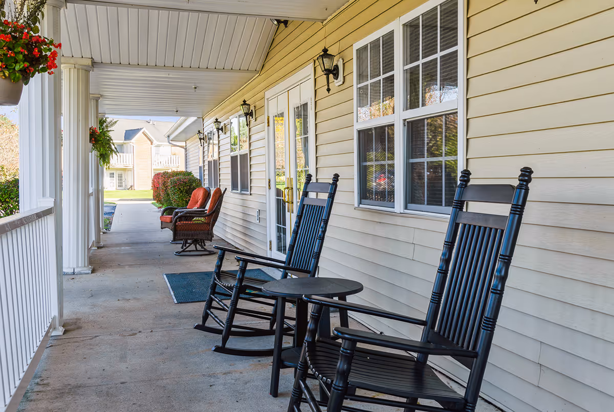 A covered porch area with two black rocking chairs and a small round table in the foreground. Further down the porch, there are two cushioned chairs with orange cushions. The porch has white railings and columns, beige siding on the building, and several wall-mounted lantern-style lights. In the background, there is a view of a lawn and neighboring buildings.