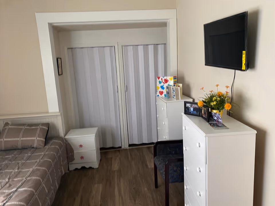 A bedroom with a bed covered in a brown checkered bedspread, a white nightstand with pink flower handles, two white dressers with various items on top including a vase with yellow and orange flowers, greeting cards, and framed photos. A flat-screen TV is mounted on the wall above one dresser. There is a chair with a dark floral cushion in front of the dressers, and sliding closet doors with vertical blinds in the background.