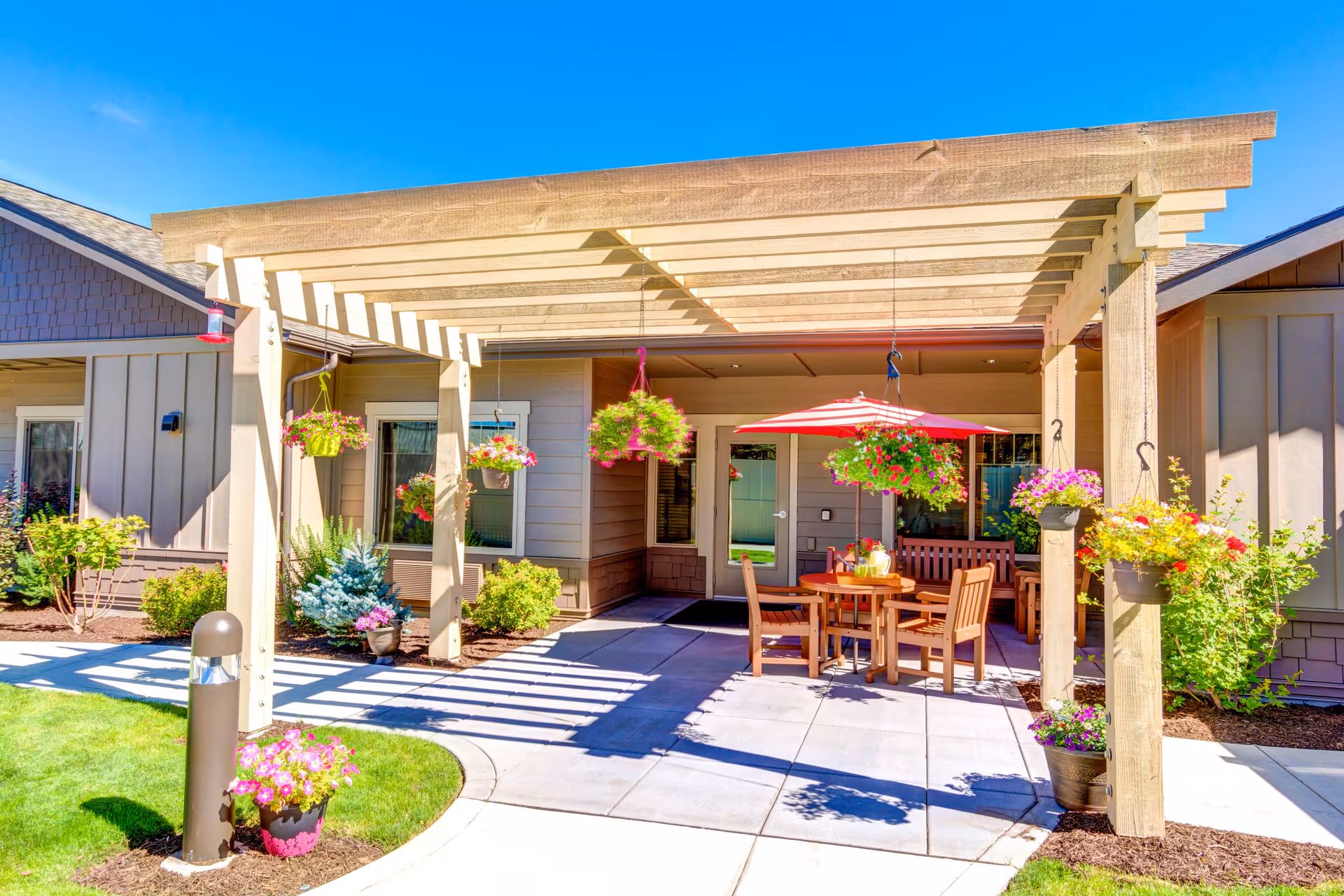 Outdoor patio area at Mt. Bachelor Assisted Living and Memory Care featuring a wooden pergola with hanging flower baskets, a round wooden table with chairs, and a red and white striped umbrella. The patio is surrounded by well-maintained landscaping with green grass, shrubs, and colorful flowers under a clear blue sky.