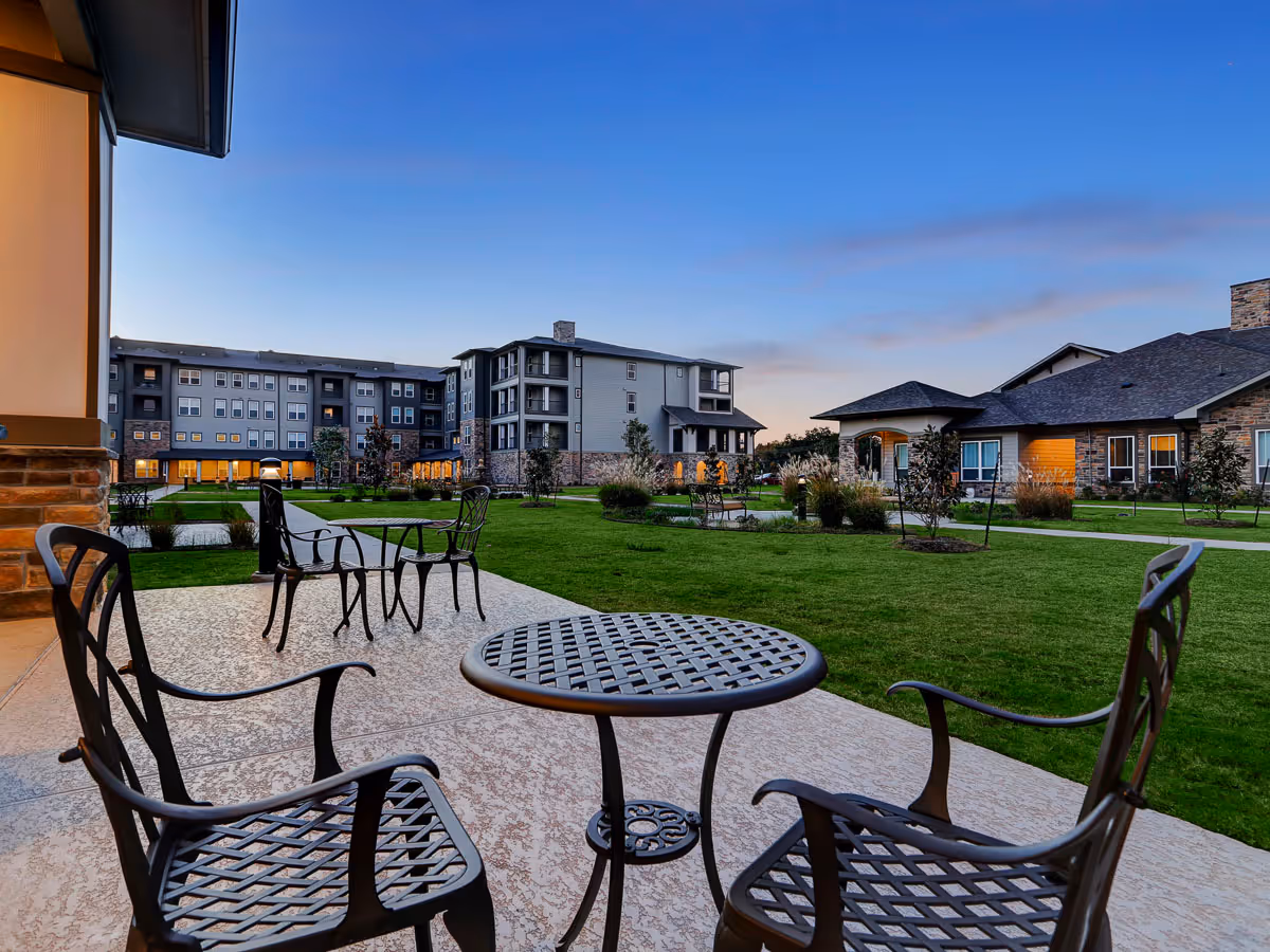 Outdoor patio area with metal tables and chairs overlooking a large grassy courtyard surrounded by multi-story and single-story buildings under a clear evening sky.