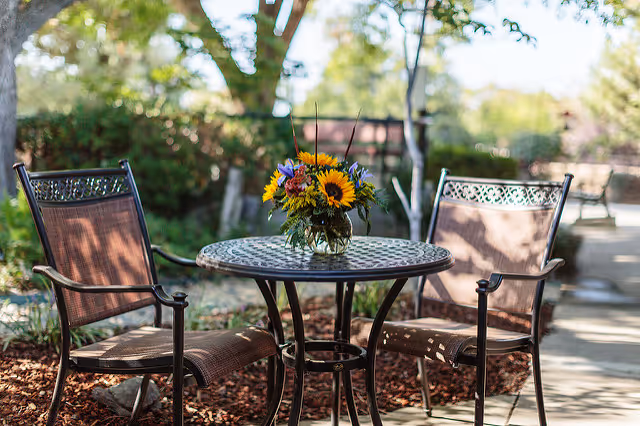 A round metal patio table holding a vase of sunflowers between two metal chairs on a shaded garden walkway.