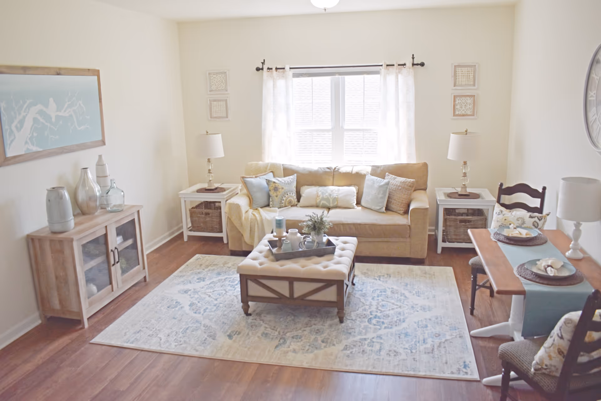 A cozy living room with a beige sofa adorned with multiple decorative pillows, two white side tables each with a lamp, a tufted ottoman coffee table with decorative items on a tray, a wooden cabinet with vases, a large window with sheer curtains, a wall clock, and a small dining table set for two with plates and napkins.