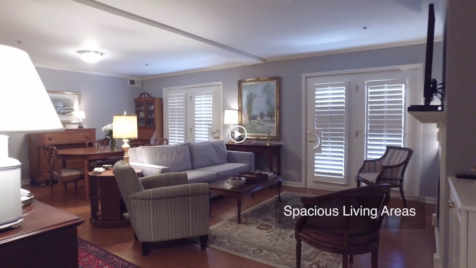 A spacious living room with a light gray sofa, two armchairs, a wooden coffee table, and a patterned area rug. The room features wooden flooring, a dining table with chairs in the background, a sideboard, and a painting on the wall. French doors with white plantation shutters allow natural light into the room.