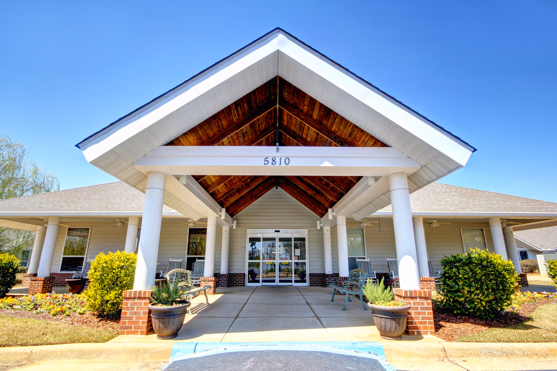 Front entrance of a senior living facility with a covered porch supported by white columns and a wooden ceiling. There are benches and potted plants on either side of the walkway leading to glass double doors. The building number 5810 is displayed above the entrance. The sky is clear and blue.