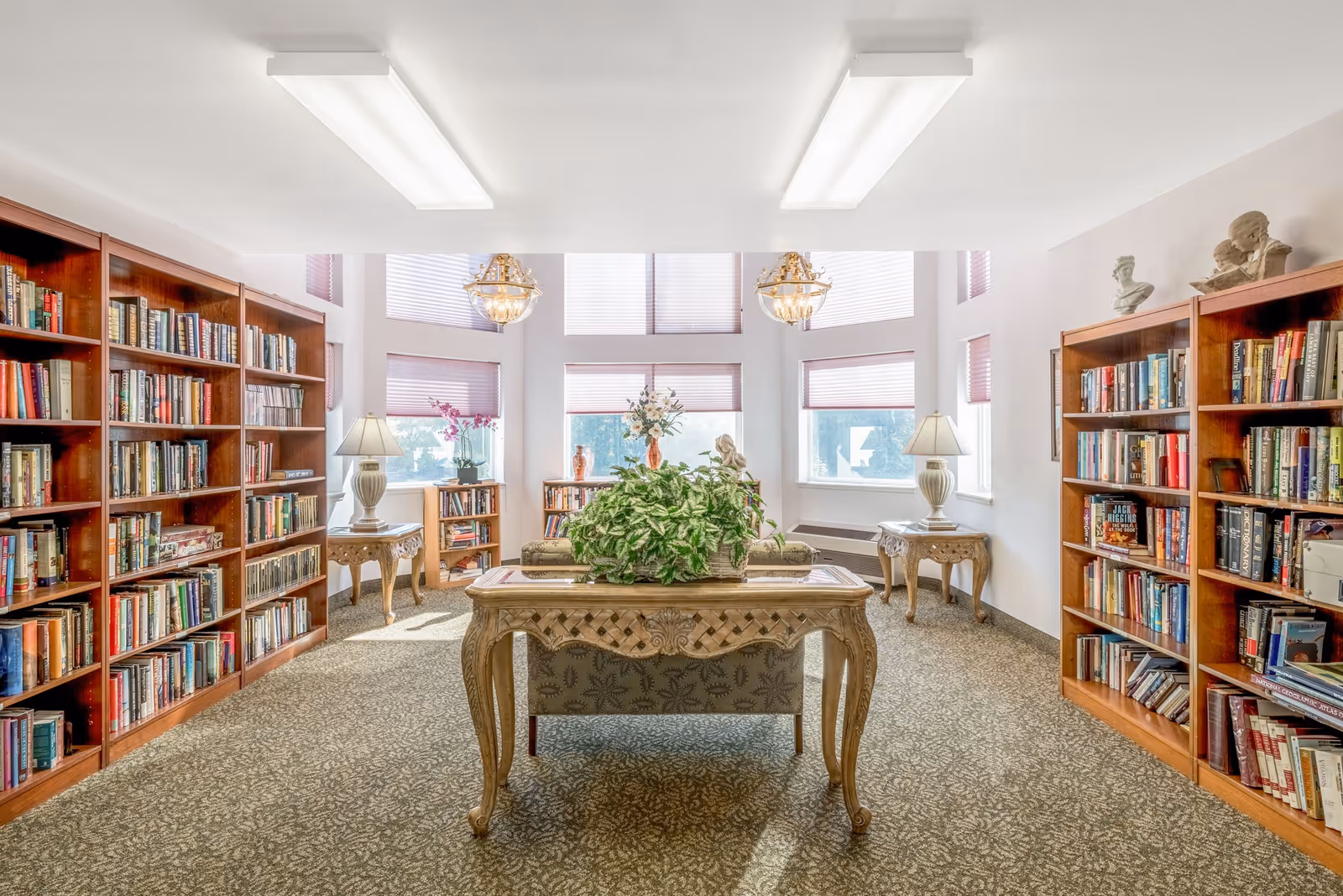A bright and spacious library room with large windows letting in natural light. The room features wooden bookshelves filled with books on both sides, two ornate table lamps on side tables, a central wooden table with a green leafy plant, and decorative busts on top of the bookshelves. The carpet has a patterned design and the ceiling has two rectangular fluorescent light fixtures.