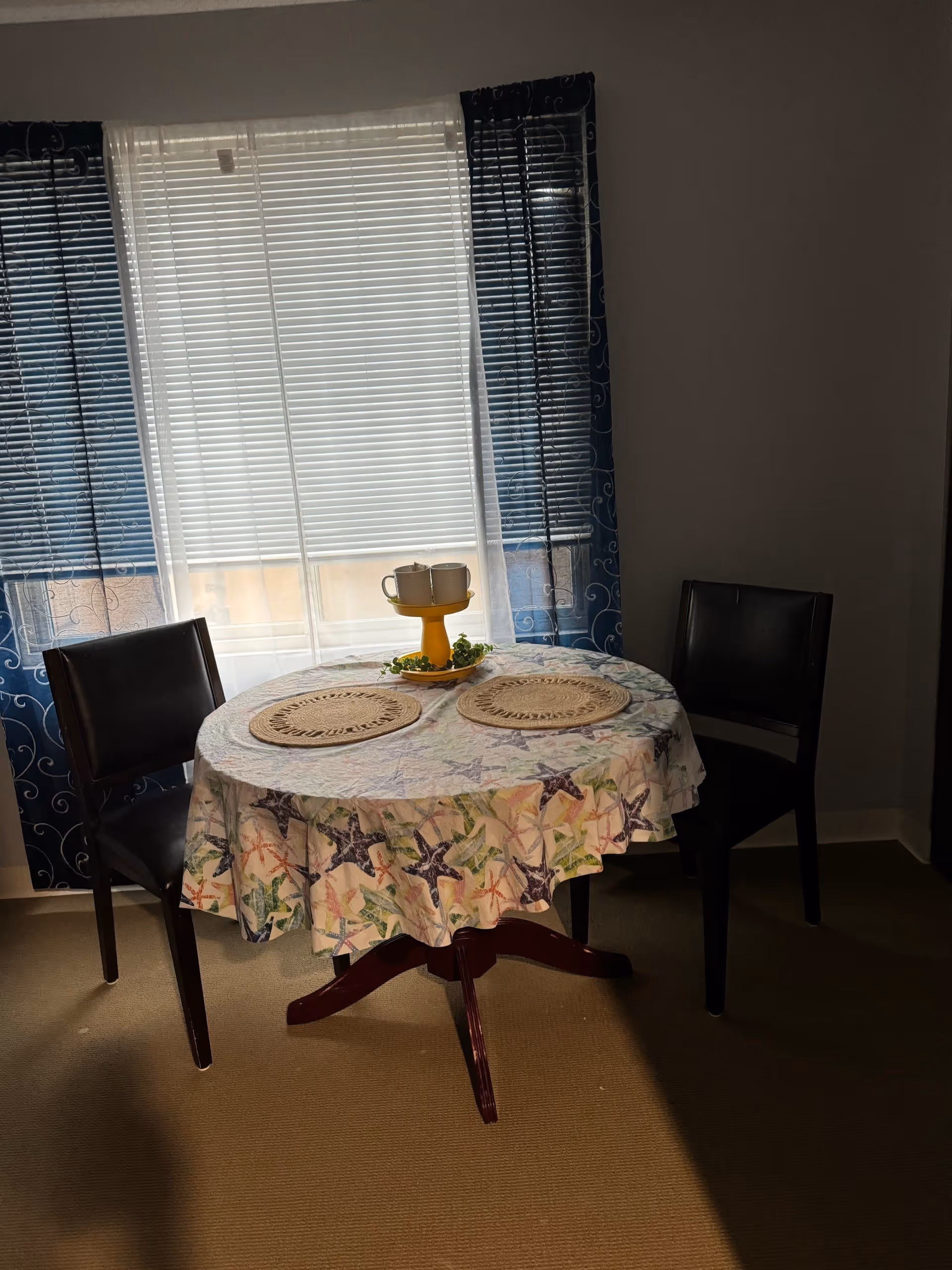 A small round dining table covered with a starfish patterned tablecloth, set with two woven placemats and a yellow two-tiered tray holding two white mugs and some greenery. The table is flanked by two dark brown chairs and positioned in front of a window with white blinds and blue curtains with a swirl pattern.