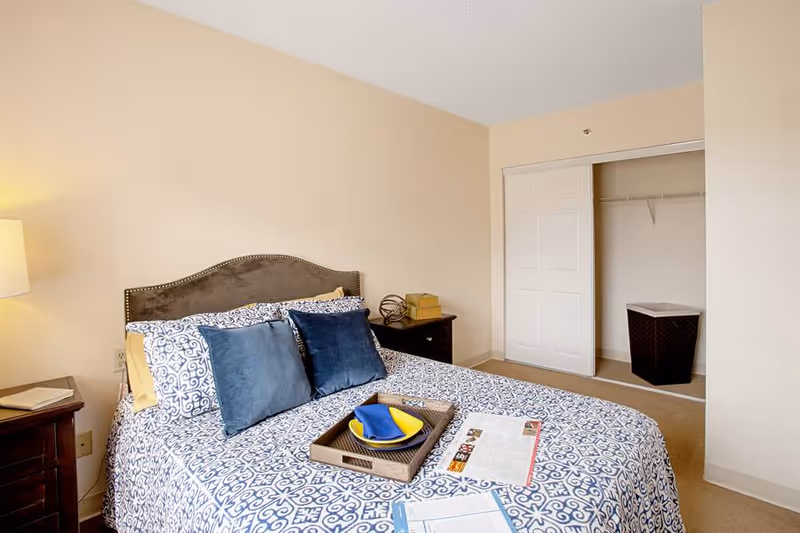 A bedroom with a bed covered in a blue and white patterned bedspread, two blue pillows, and a tray with a yellow plate and blue napkin. There is a nightstand with a lamp and a book on the left side of the bed, and another nightstand with decorative items on the right. In the background, there is an open closet with a laundry basket inside.